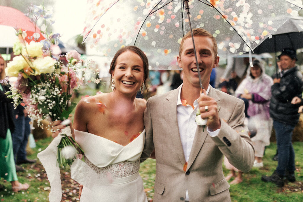 The newly weds walk hand-in-hand down the aisle as husband and wife with colourful confetti falling onto them. Happy wedding portrait captured by Eilish Burt Photography