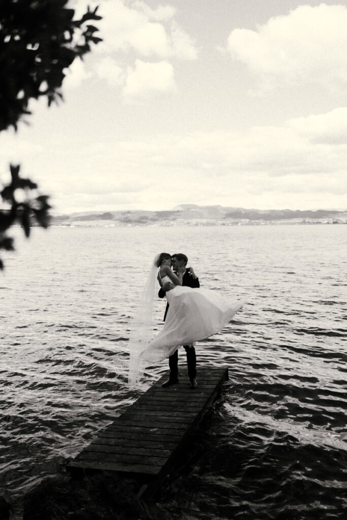 A wedding portrait of a Husband holding his new wife on the edge of a jetty at Peppers On The Point in Rotorua. Captured by Eilish Burt Photography