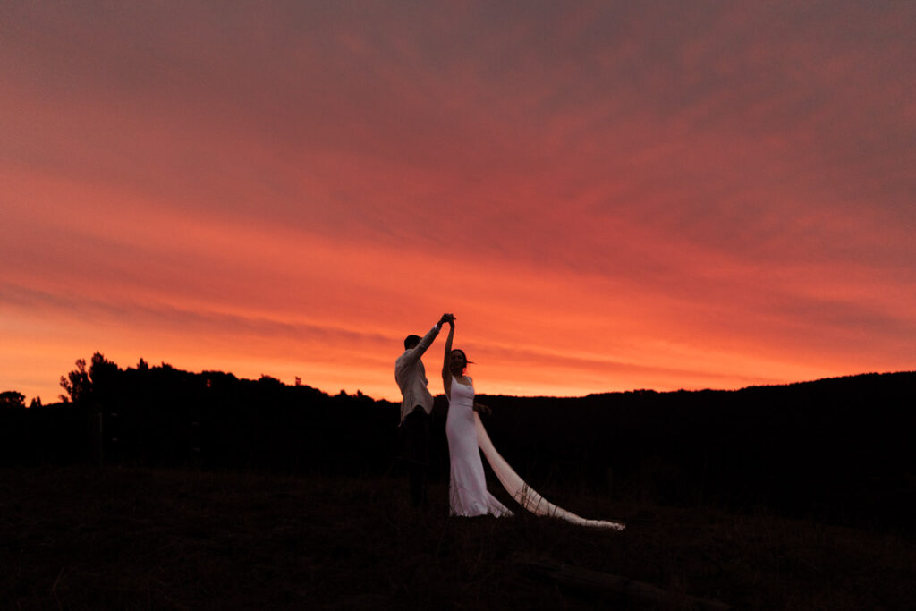 The newly married couple dance against a striking red sky as the sunsets. Captured by Eilish Burt Photography in Whakatane