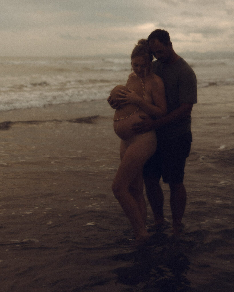 A pregnant couple lovingly hold each other at their maternity session in Ohope Beach captured by Eilish Burt Photography