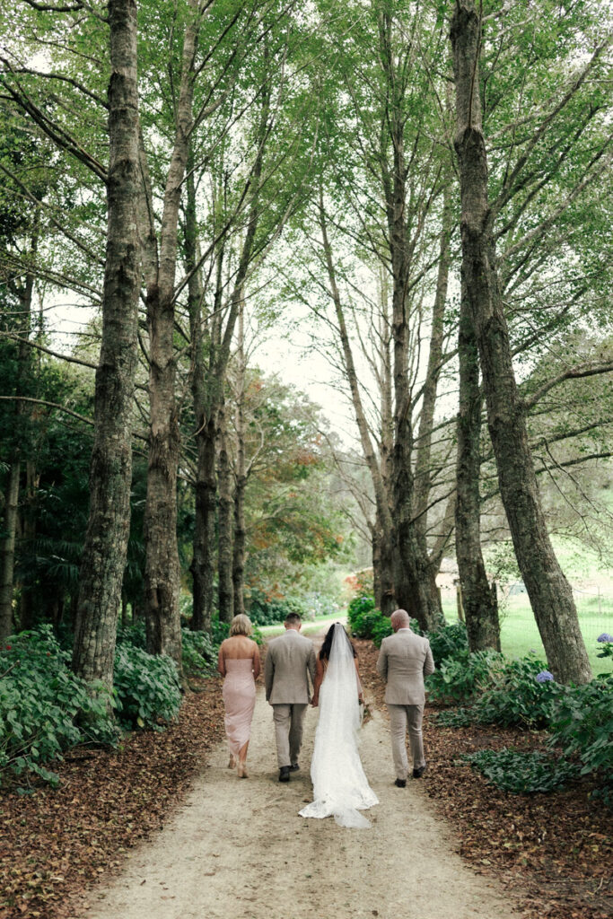 The bridal party walking down a tree covered driveway on their wedding day at The Run 15 in Whakatane. Captured by Eilish Burt Photography