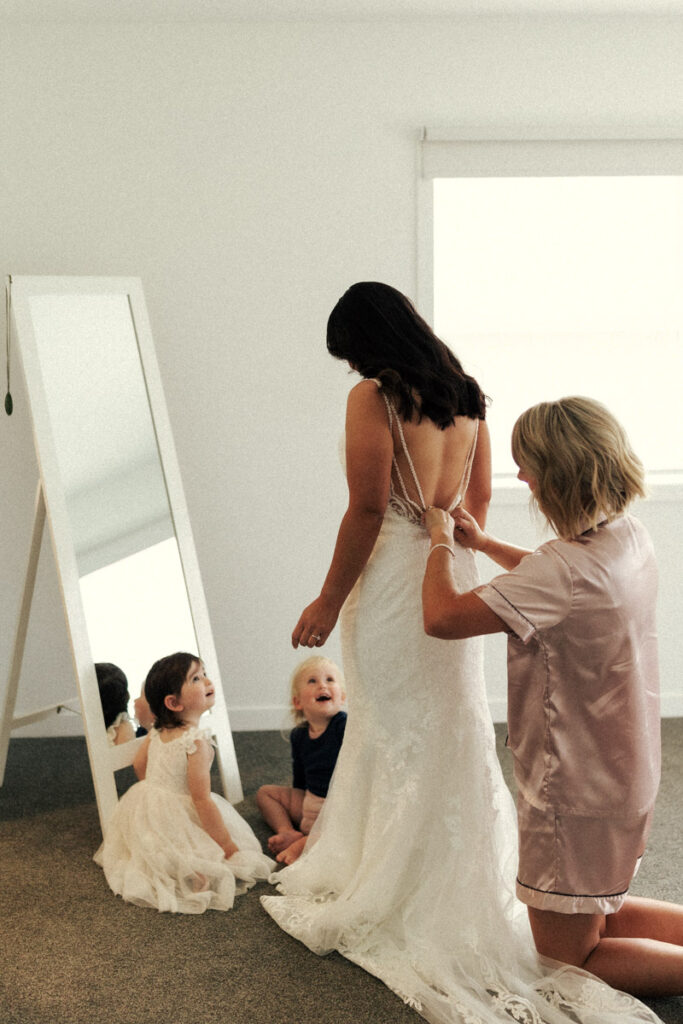 The brides daughter looks up at her as her mum is getting her wedding dress put on before she says I Do to her dad - the groom. Captured by Eilish Burt Photography