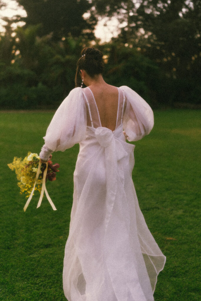 A captivating film portrait of the back of the brides dress as she walks through across the lawn at the Puketutu Island Estate. Captured by Eilish Burt Photography