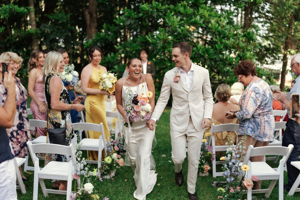 A bride and groom walk down the aisle as husband and wife amongst the vibrant coloured flowers in Byron Bay, Australia