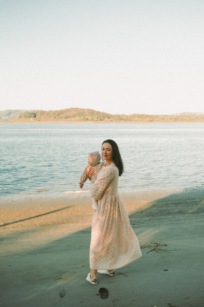 Mum stands at the harbour of Ohope while holding her baby on her hip. Photographed by Eilish Burt Photography.