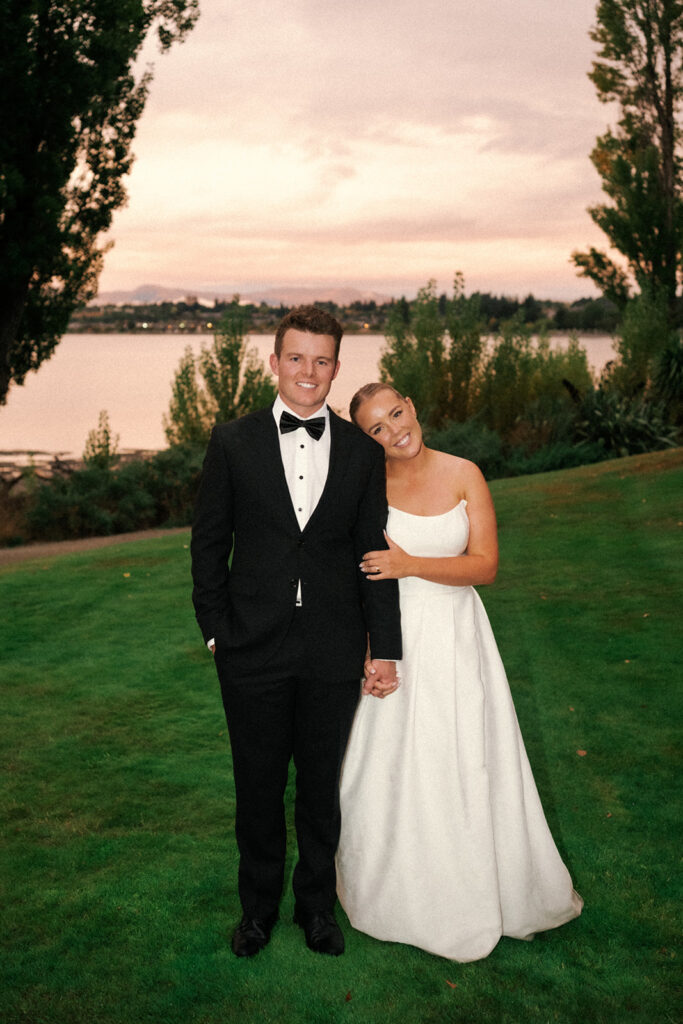 Bride and groom smile for their camera with the golden sun setting behind them at the Edgewater Estate in Wanaka.